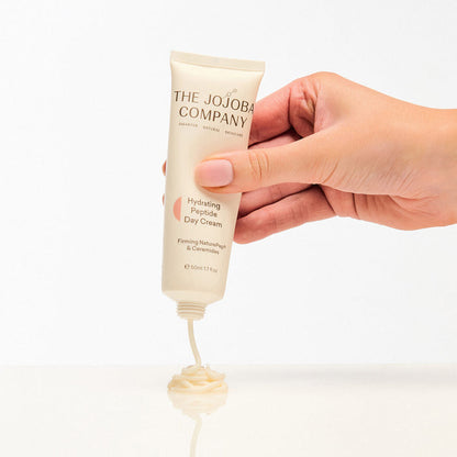 Hand holding a tube of 'The Jojoba Company' cream with visible cream at the end on a white background.