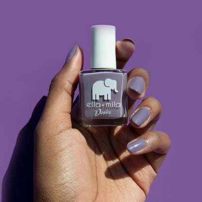 Mauve Over grey-purple nail polish on a deep skin tone against a purple background, holding the bottle