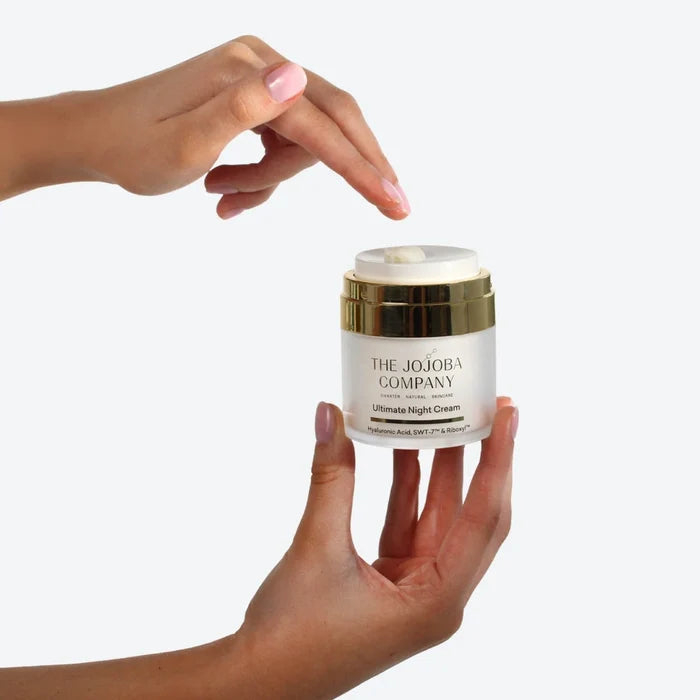 Hand holding a jar of 'The Jojoba Company' Ultimate Night Cream with some pumped into the top against a white background
